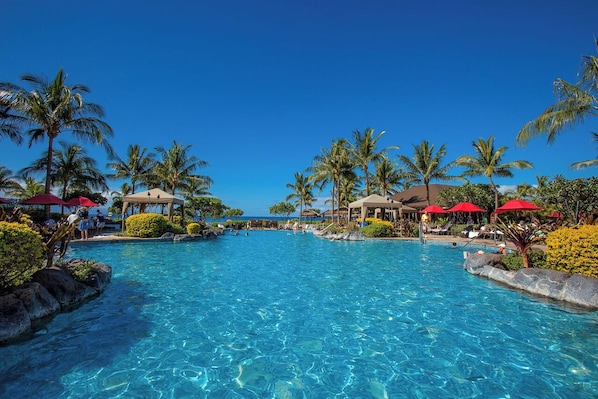 A clear blue pool surrounded by palm trees and red umbrellas, with lounge chairs and a view of the ocean.