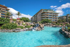 A resort pool with a water slide, surrounded by multi-story buildings with balconies, and lush greenery.