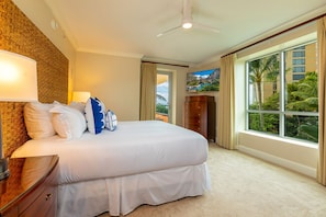 A hotel room with a large bed, a ceiling fan, a TV, and a view of palm trees through the windows.