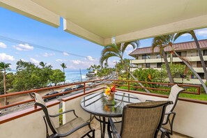 A balcony with a glass table and chairs, overlooking a coastal road and ocean.