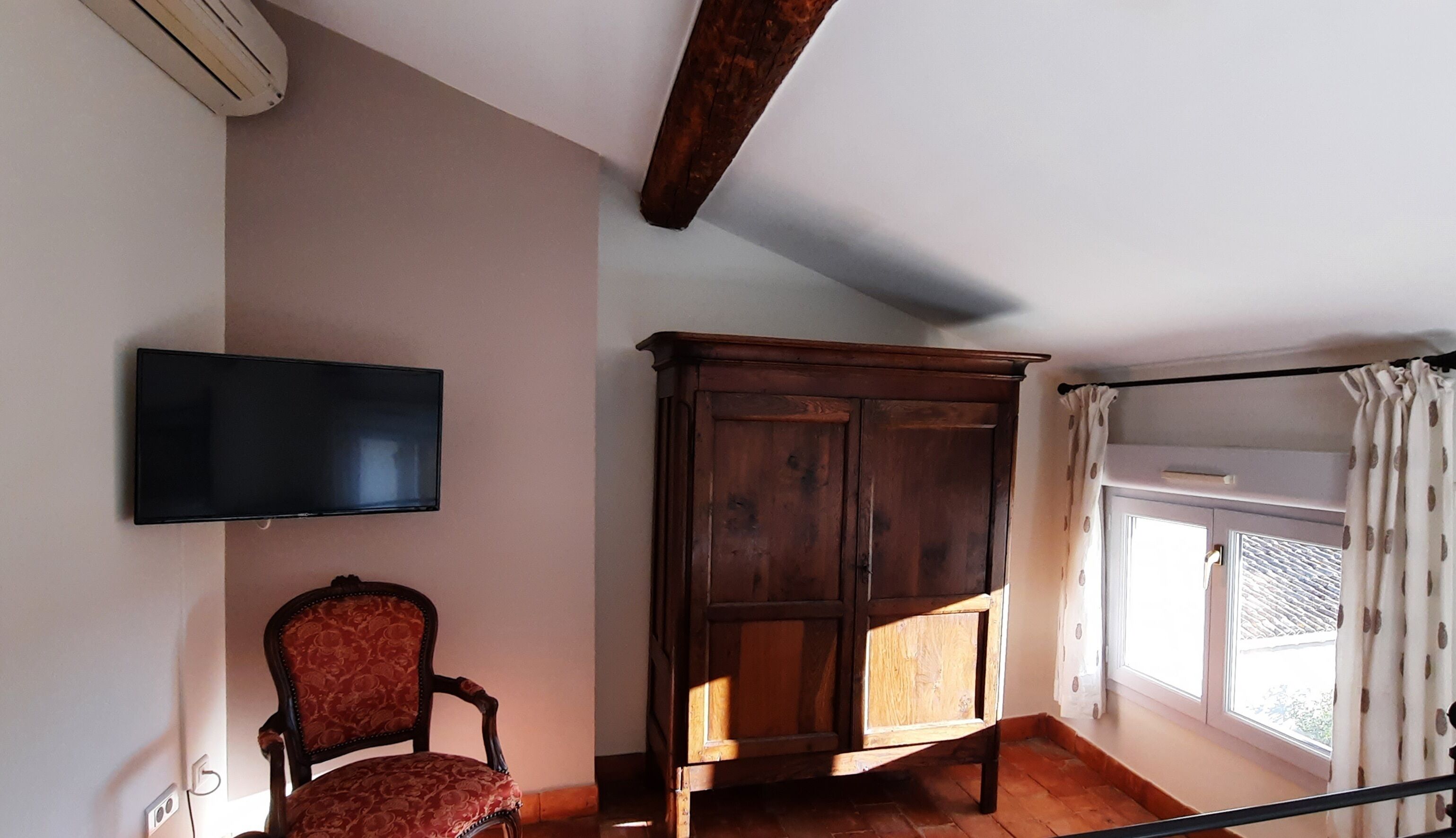 Une pièce comprenant un meuble en bois, un fauteuil rouge recouvert de tissu, un téléviseur à écran plat fixé au mur et une fenêtre avec des rideaux.