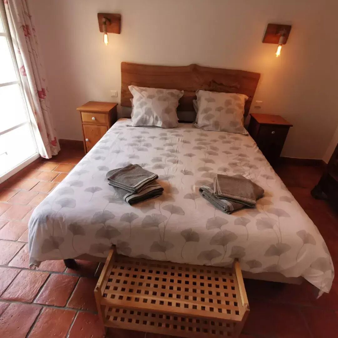 Een net opgemaakt bed met een bedsprei met patroon, twee nachtkastjes, een houten bankje en een raam met gordijnen.