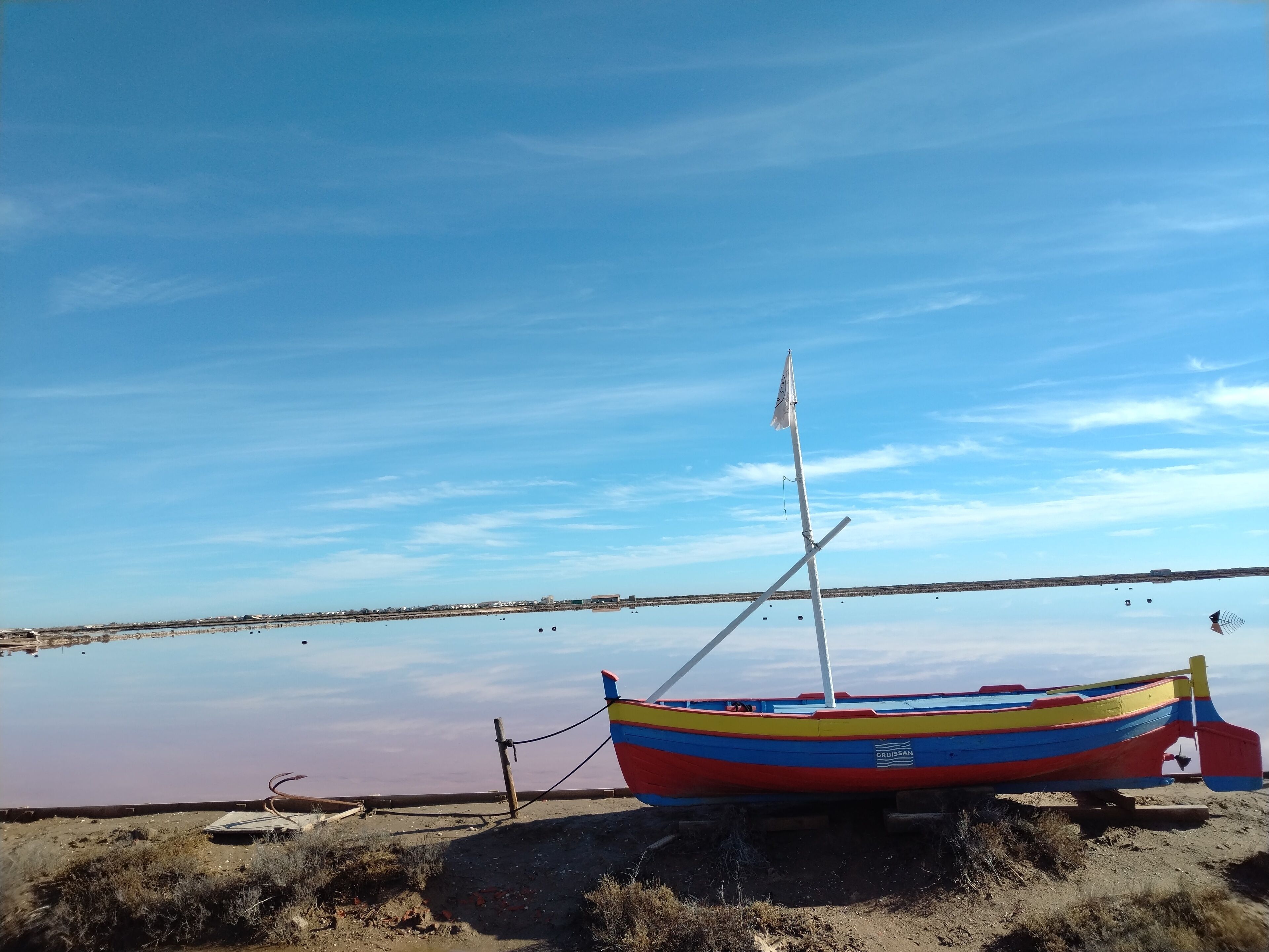 Un bote colorido con una bandera, atracado en una orilla arenosa con un cuerpo de agua tranquilo y un cielo despejado al fondo.