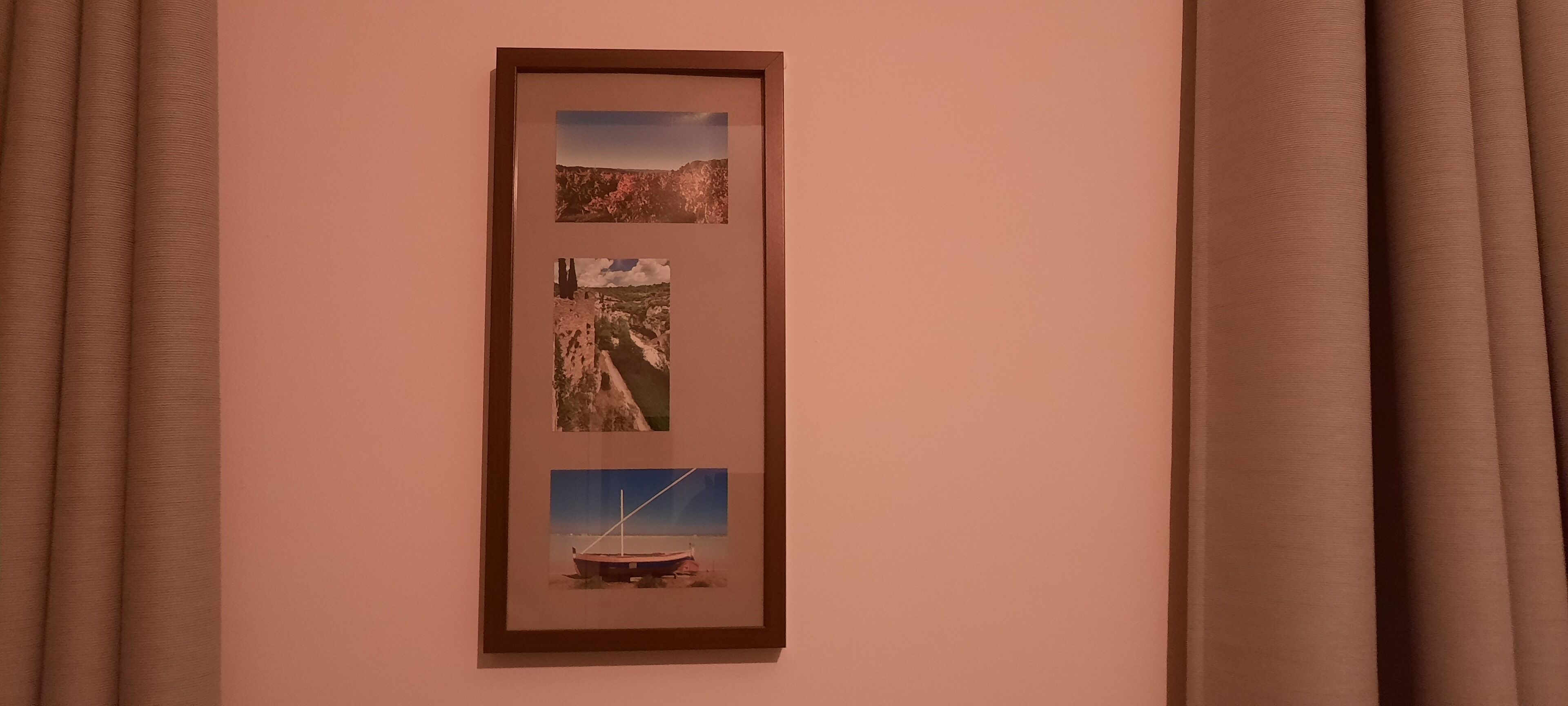 A framed photograph of a coastal landscape with cliffs and a boat on the shore, hung on a wall with brown curtains.