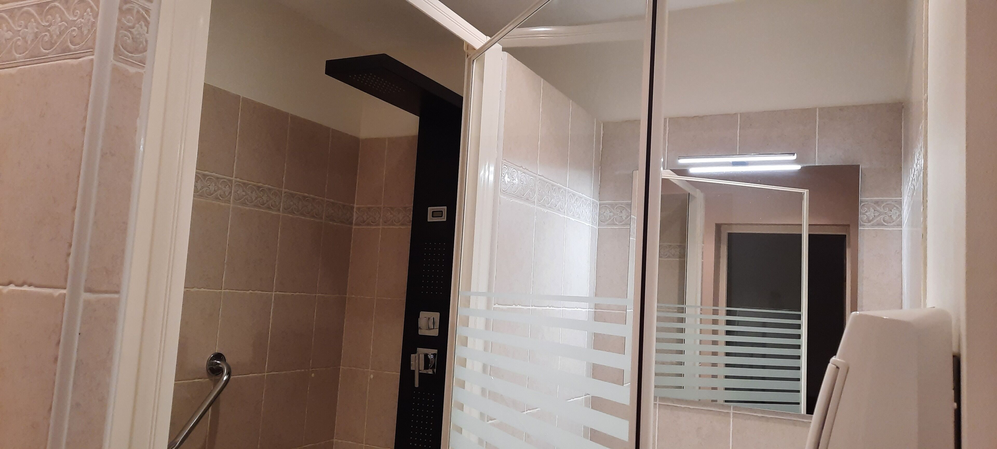 Un baño con azulejos color beige, zona de ducha, puerta de cristal con diseño texturizado y ventana con persianas.