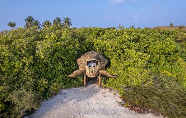 A thatched-roof hut with a table and chairs in the center, surrounded by dense greenery.