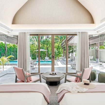 A modern hotel room with a large window, a bed, two chairs, a small table, and a view of a pool and greenery outside.