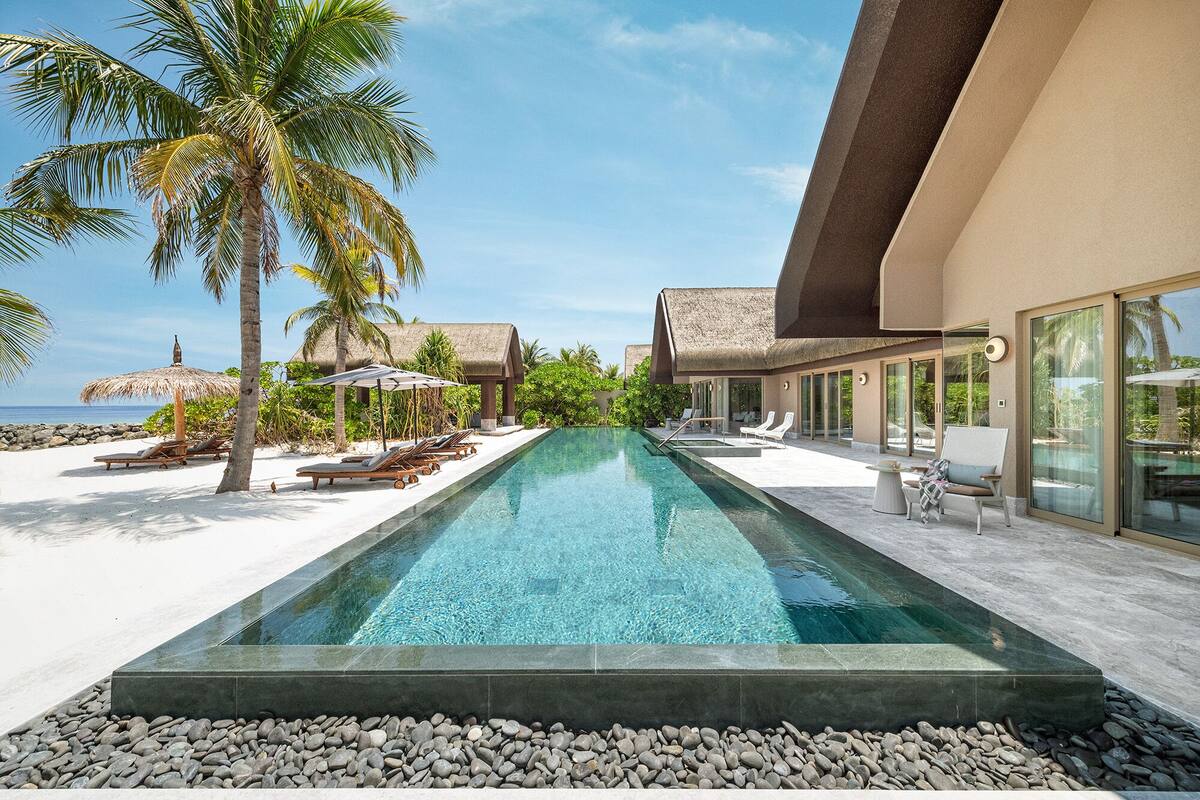 Piscina moderna con agua cristalina, rodeada de tumbonas y palmeras, que conduce a un edificio con grandes ventanales y una estructura de techo de paja.
