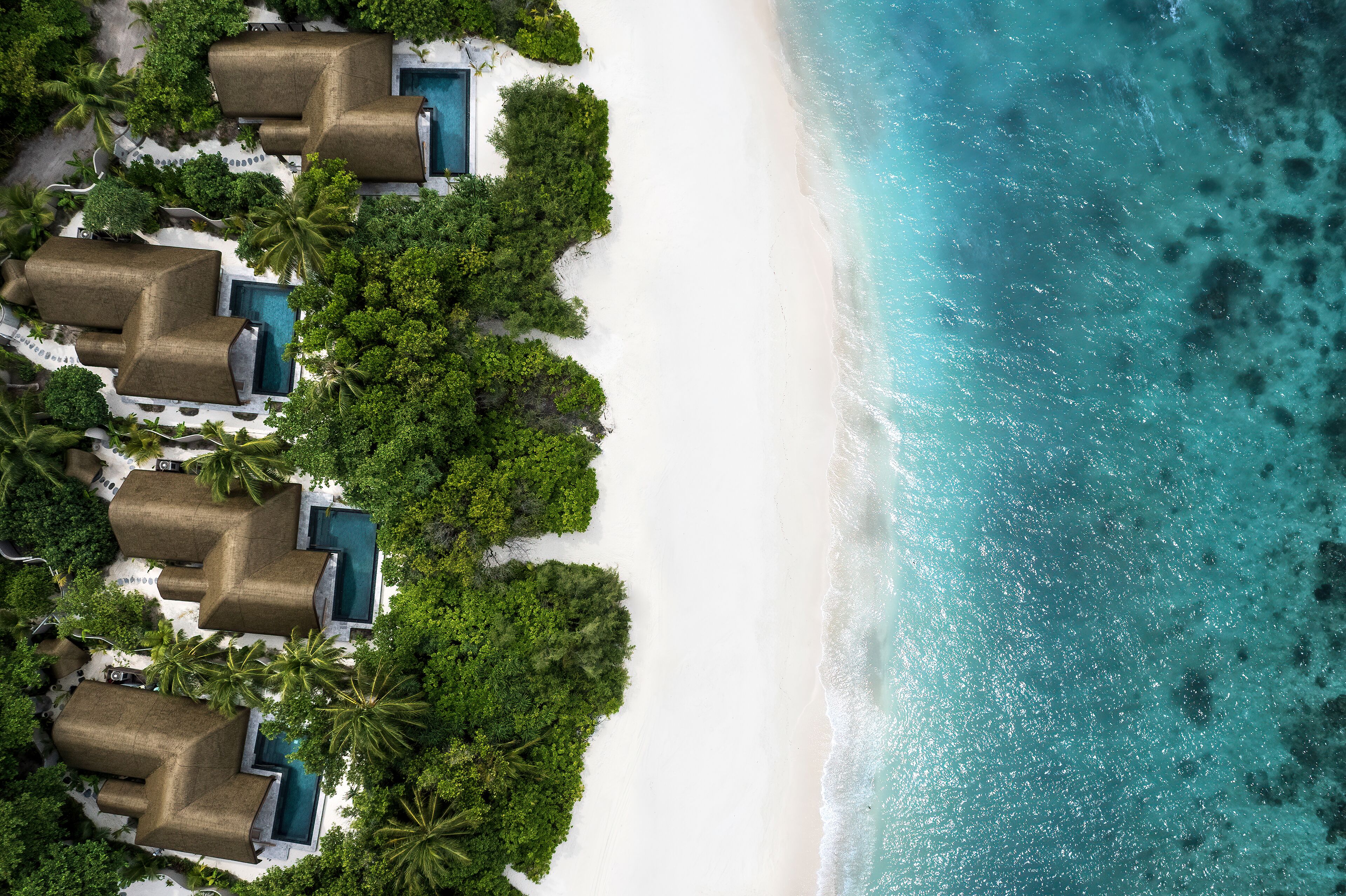 An aerial view of a beach resort with multiple overwater bungalows, each with a private pool, surrounded by lush greenery and white sand.