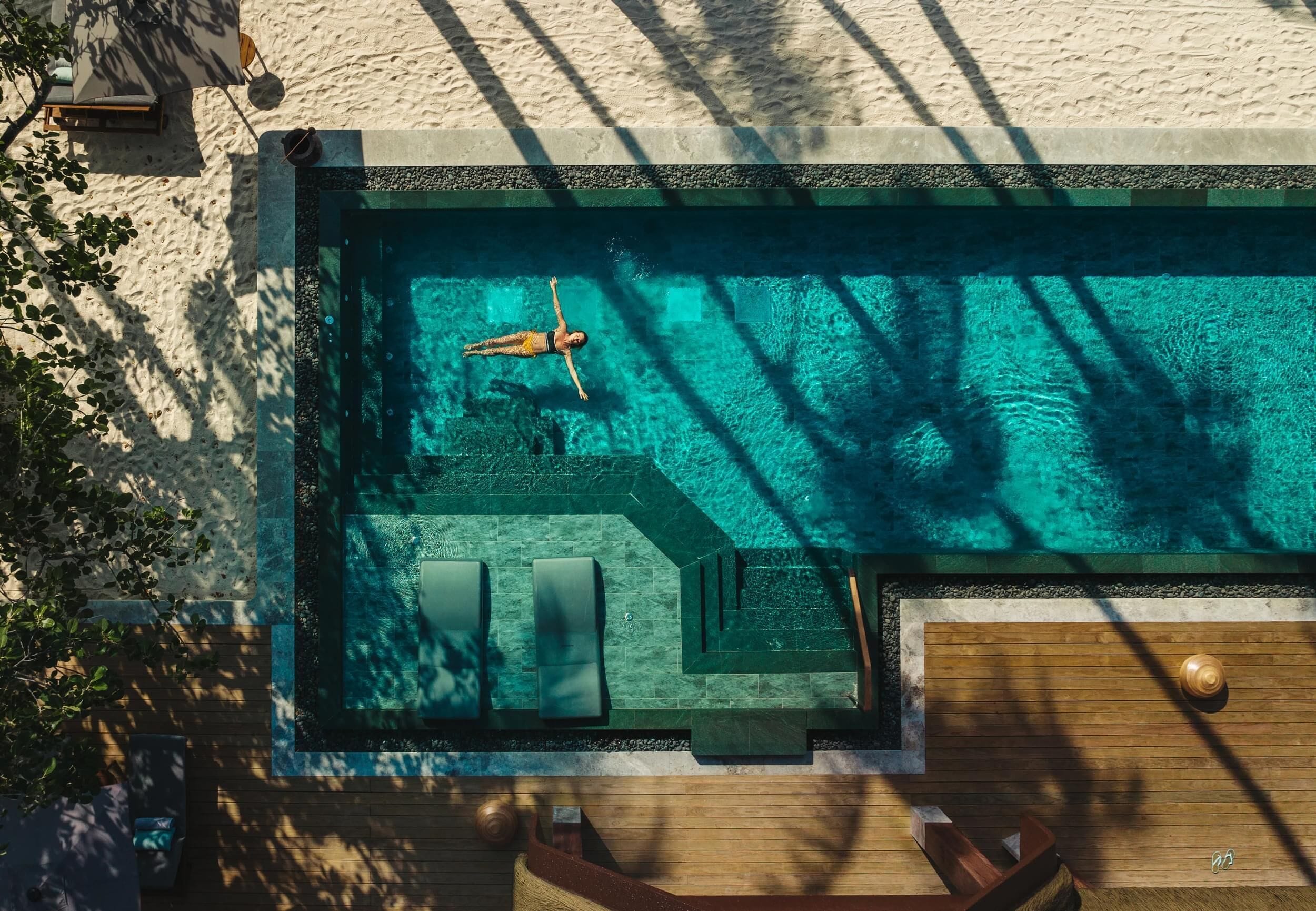 An aerial view of a swimming pool with a person swimming, surrounded by lounge chairs and a wooden deck.