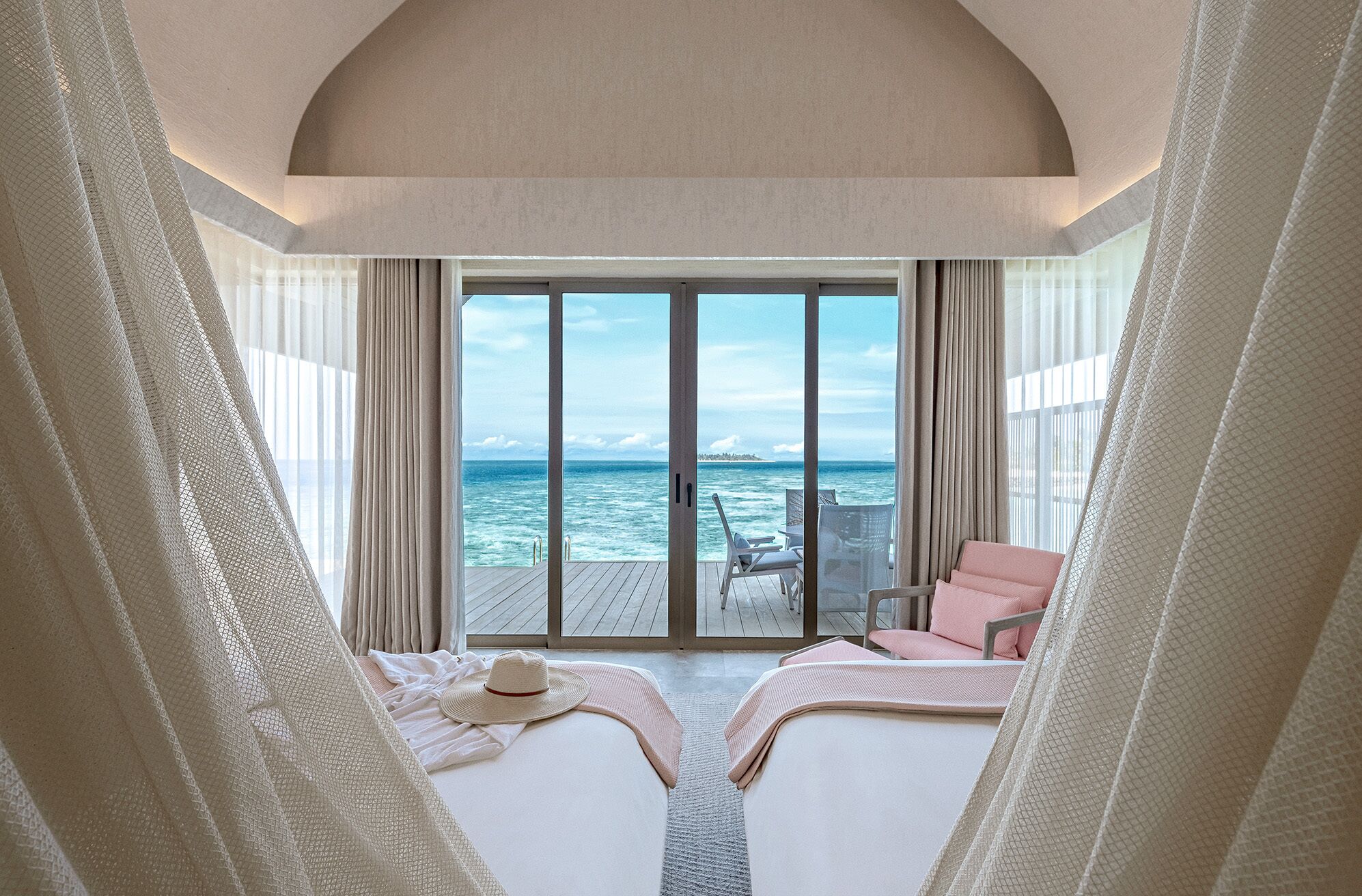 A room with a large sliding glass door leading to a deck with a view of the ocean. There are two beds with white linens and a pink chair.