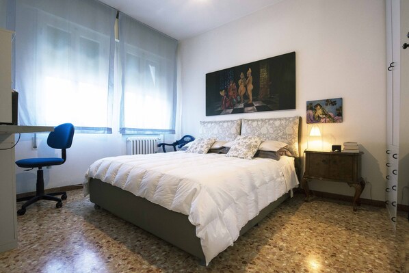 A bedroom with a bed, a desk, a chair, a radiator, and a window with curtains.