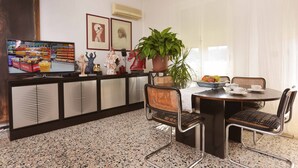 Zona pranzo con tavolo rotondo, quattro sedie, una pianta in vaso e un televisore che mostra un supermercato.