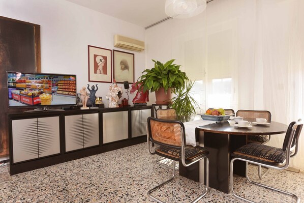 A dining area with a round table, chairs, a television displaying a supermarket, and a potted plant.