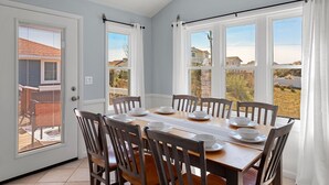 Un coin repas avec une table dressée pour six personnes, six chaises en bois et de grandes fenêtres ornées de rideaux blancs.