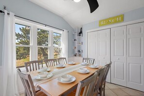 Un coin repas avec une table en bois dressée pour six personnes, des chaises blanches et une vue sur l’extérieur grâce à de grandes fenêtres.