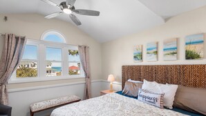 Une chambre à coucher comprenant un lit, un banc, une table de chevet avec une lampe, un ventilateur de plafond et une fenêtre avec des rideaux.