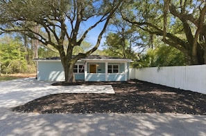 A small, single-story house with a porch, a white picket fence, and a large tree in front.
