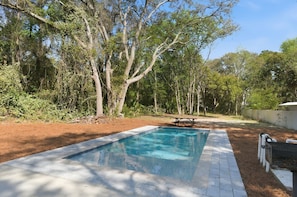 A rectangular swimming pool with a tiled deck, surrounded by trees and a picnic table.