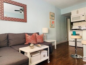 A modern living room with a grey sofa, a white side table, and a red patterned throw pillow. There is a small white table with a lamp and a decorative bowl. A kitchen area with a white microwave and cabinets is visible in the background.