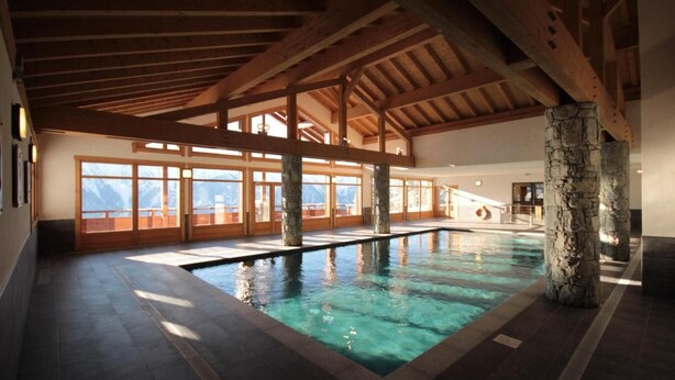 An indoor swimming pool with stone pillars, wooden beams, and large windows offering mountain views. - 100m from ski slopes, 3rd floor, view panoramic, swimming pool, balcony, parking, tv, ski locker (Villard-sur-Doron)