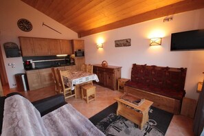 Ein gemütliches Wohnzimmer mit einer Holzdavonia, einem Esstisch mit Stühlen, einer Küchenzeile mit Holzschränken und einem an der Wand montierten Flachbildfernseher.