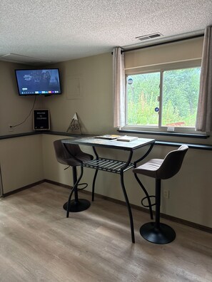 A room with a desk, two chairs, a television mounted on the wall, a window with curtains, and a welcome sign.