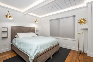 A bedroom with a wooden bed, white walls, a large window with blinds, and a floor lamp.