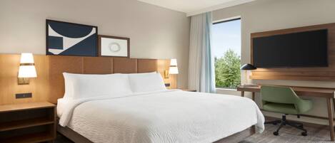Una habitación de hotel con una cama grande, dos mesitas de noche con lámparas, un escritorio con una silla, un televisor de pantalla plana y una ventana con vistas a árboles.
