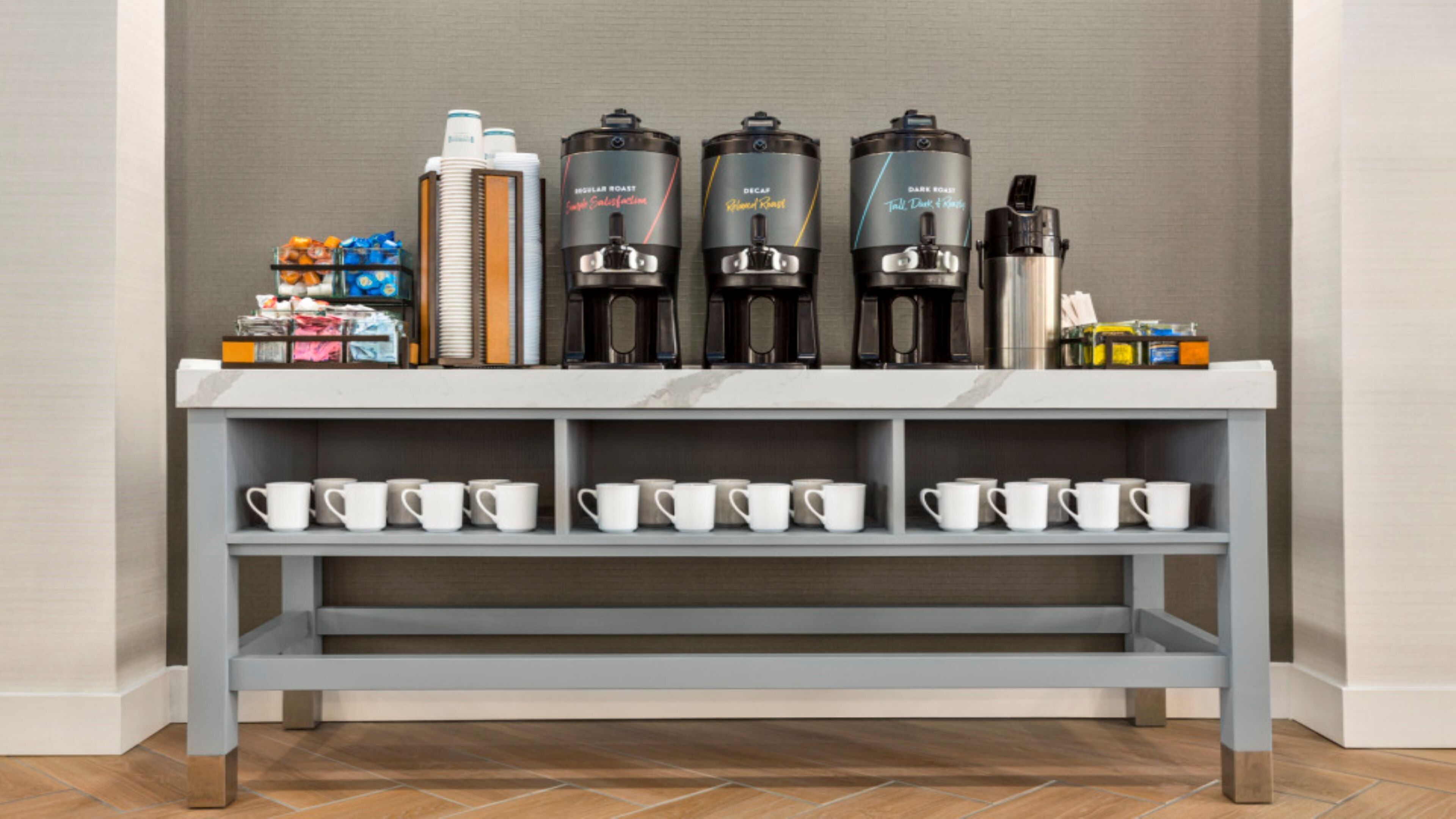 A coffee station with four coffee makers, a stack of disposable cups, and a shelf with white mugs.