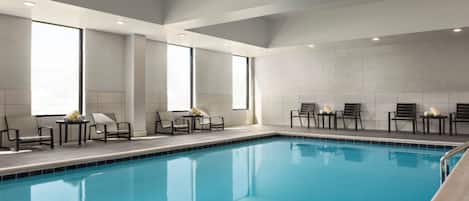 Una piscina interior con sillones y mesas, ventanas amplias y un muro de azulejos.