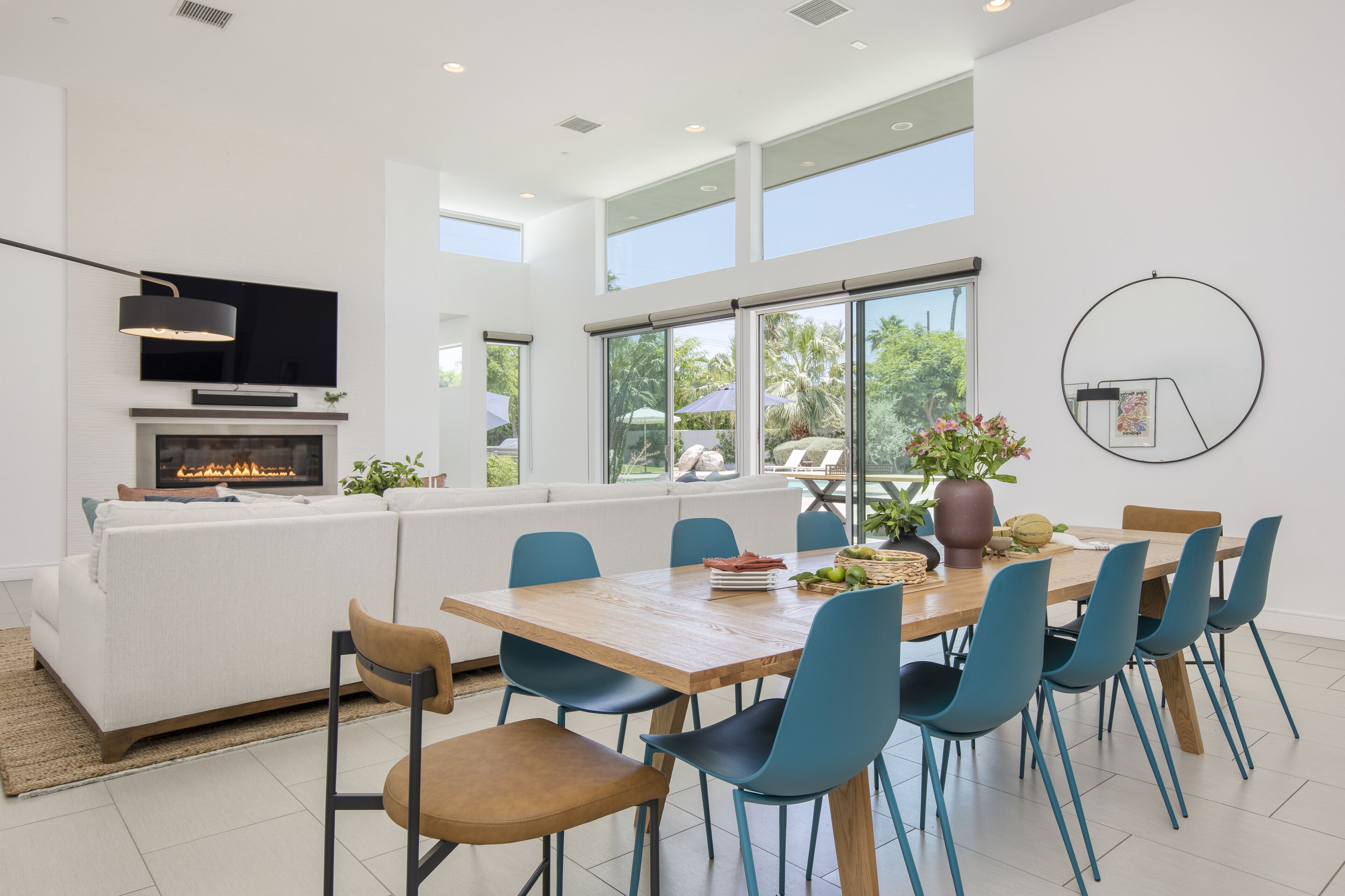 A modern living room with a fireplace, a dining area with blue chairs, and large windows offering a view of the outdoors.