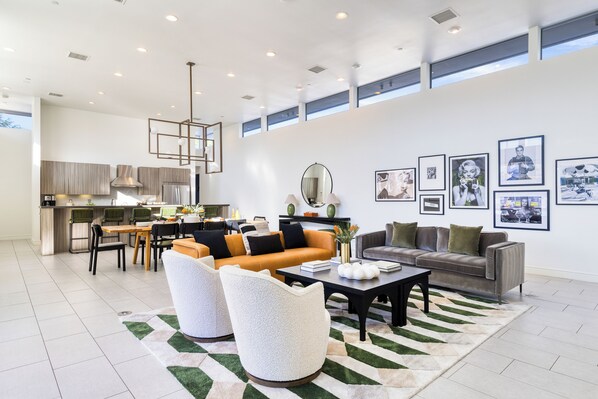 Una sala de estar moderna con sofá, mesa de centro y sillones. Se ve una cocina al fondo con una mesa de comedor y sillas.