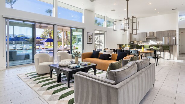 A spacious living area with a sofa, armchairs, and a coffee table. A kitchen is visible in the background with modern appliances. Large windows provide natural light and a view of the outdoors.