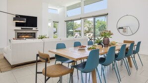 Un salon moderne avec une cheminée, un coin repas avec des chaises bleues et de grandes fenêtres donnant sur l’extérieur.
