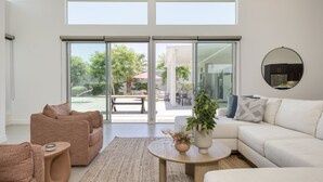 Un vaste salon doté d’une grande porte-fenêtre coulissante donnant sur une terrasse extérieure, d’un canapé blanc, d’un fauteuil beige et d’une table basse sur laquelle est posé un vase de fleurs.