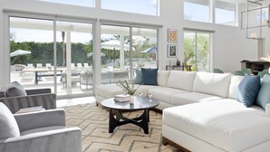 Ein geräumiges Wohnzimmer mit einem weißen Sofa, einem runden Couchtisch und einer großen Glasschiebetür, die zu einem Außenbereich mit Pool und Sonnenschirmen führt.