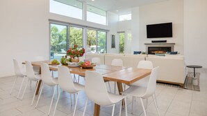 A modern dining area with a wooden table, white chairs, and a large window offering a view of greenery.