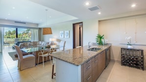 Una cocina moderna con una isla central, un comedor con mesa de vidrio y vista a palmeras al exterior.