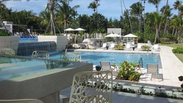 Un área de piscina con sillones de descanso y sombrillas, rodeada de palmeras.