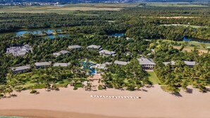Vista aérea de un resort con varias edificaciones, una playa y una zona de piscina rodeada de palmeras.