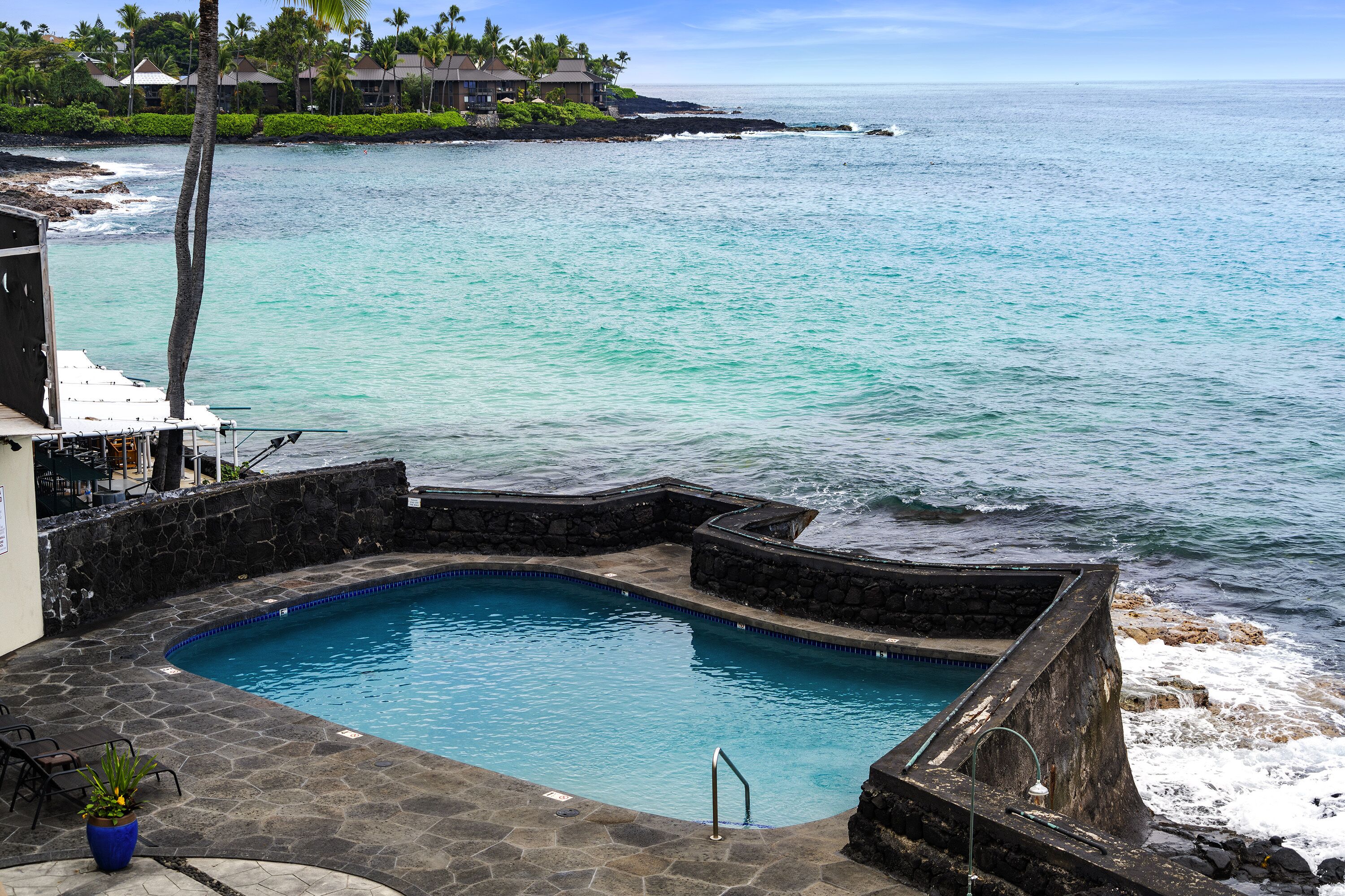 A pool with a stone border, surrounded by a stone deck, overlooking a beach with a view of the ocean and a distant shoreline with buildings.