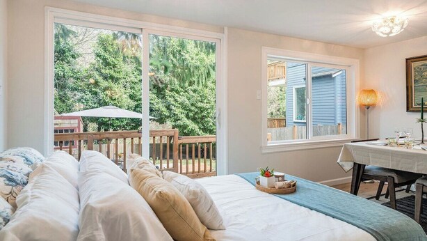 A bedroom with a large bed, a dining table with chairs, and a view of a wooden deck and trees. - Perch - Brygger Cottage: Magnolia rambler & pet-friendly (Seattle)