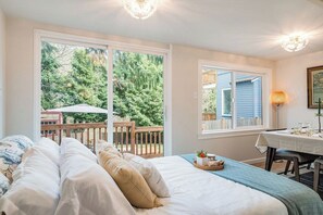 Une chambre avec un grand lit, une table à manger avec des chaises, et une vue sur une terrasse en bois et des arbres.