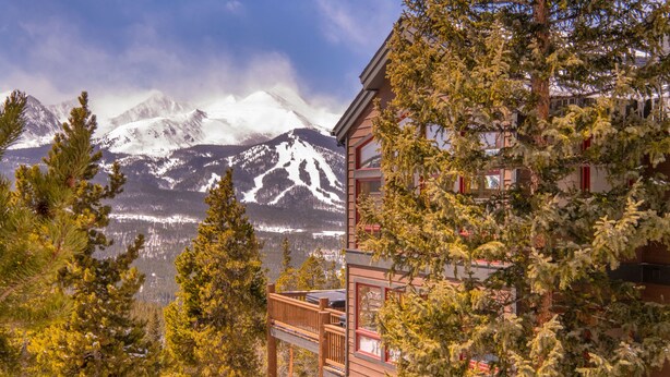 Un edificio de varios pisos con un balcón, rodeado de árboles y con vistas a una cordillera nevada.