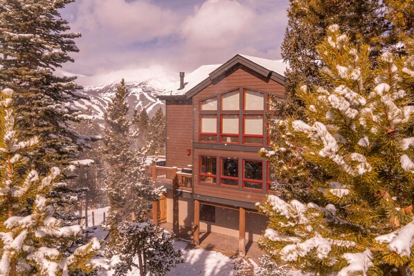 Eine zweistöckige Holzhütte mit Balkon, umgeben von schneebedeckten Bäumen und Bergen im Hintergrund.