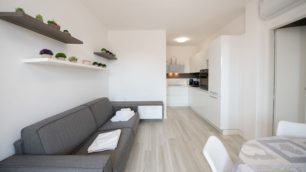 Ein modernes Wohnzimmer mit einem grauen Sofa, einem Esstisch mit gemustertem Tischläufer und weißen Stühlen. Im Hintergrund sind weiße Schränke und eine Küchenzeile zu sehen.