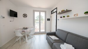 Ein modernes Wohnzimmer mit grauem Sofa, einem Esstisch mit Stühlen, einem flachbildfernseher an der Wand und einem großen Fenster mit Schiebetür.