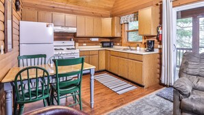 A cozy cabin-style kitchen with wooden cabinets, a stove, a microwave, and a coffee maker. There is a dining table with chairs and a sofa in the living area.