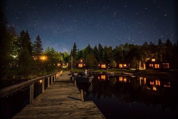 A wooden dock extending into a calm body of water with houses on the opposite shore, under a starry night sky.